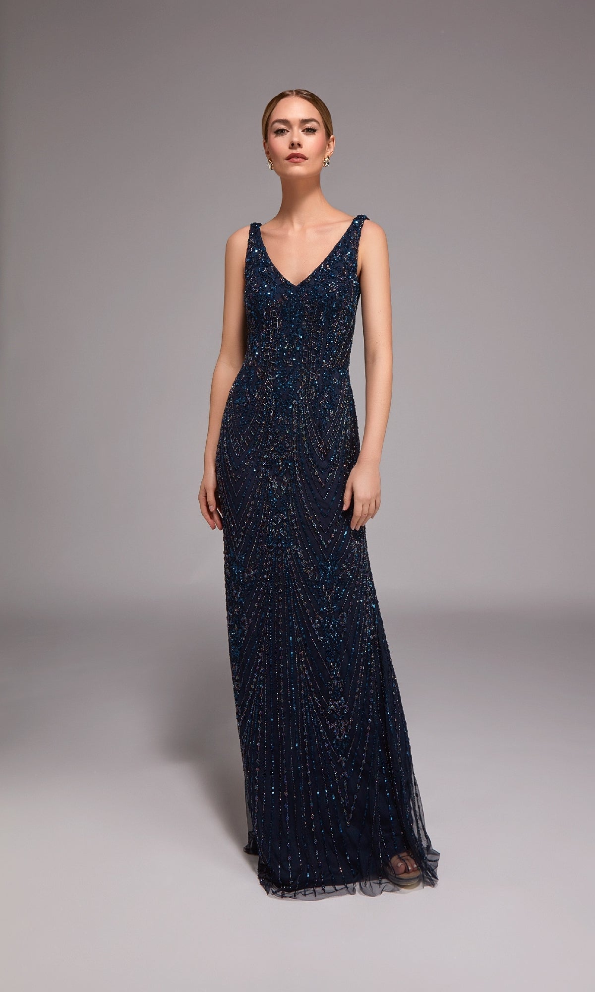 A woman poses in a studio wearing the Alyce 27786, a long formal sleeveless column gown with beading and V-neckline, perfect as an elegant evening dress in deep navy blue.